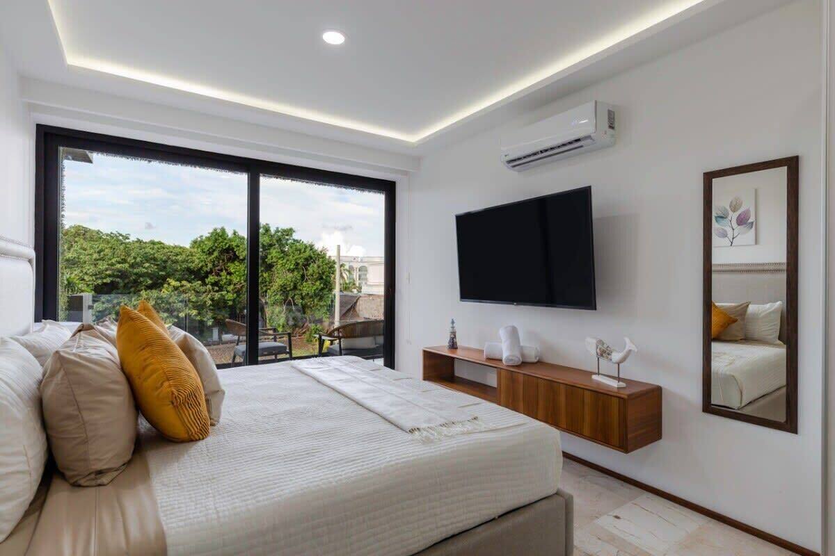 A modern bedroom with a large bed, a flat-screen TV, a bedside table, and a view of greenery outside.