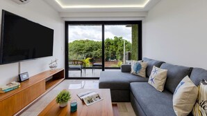 Een moderne woonkamer met een flatscreen televisie, een grijze bank met decoratieve kussens, een houten salontafel met een plant, en een schuifpui die leidt naar een balkon met buitenmeubilair.