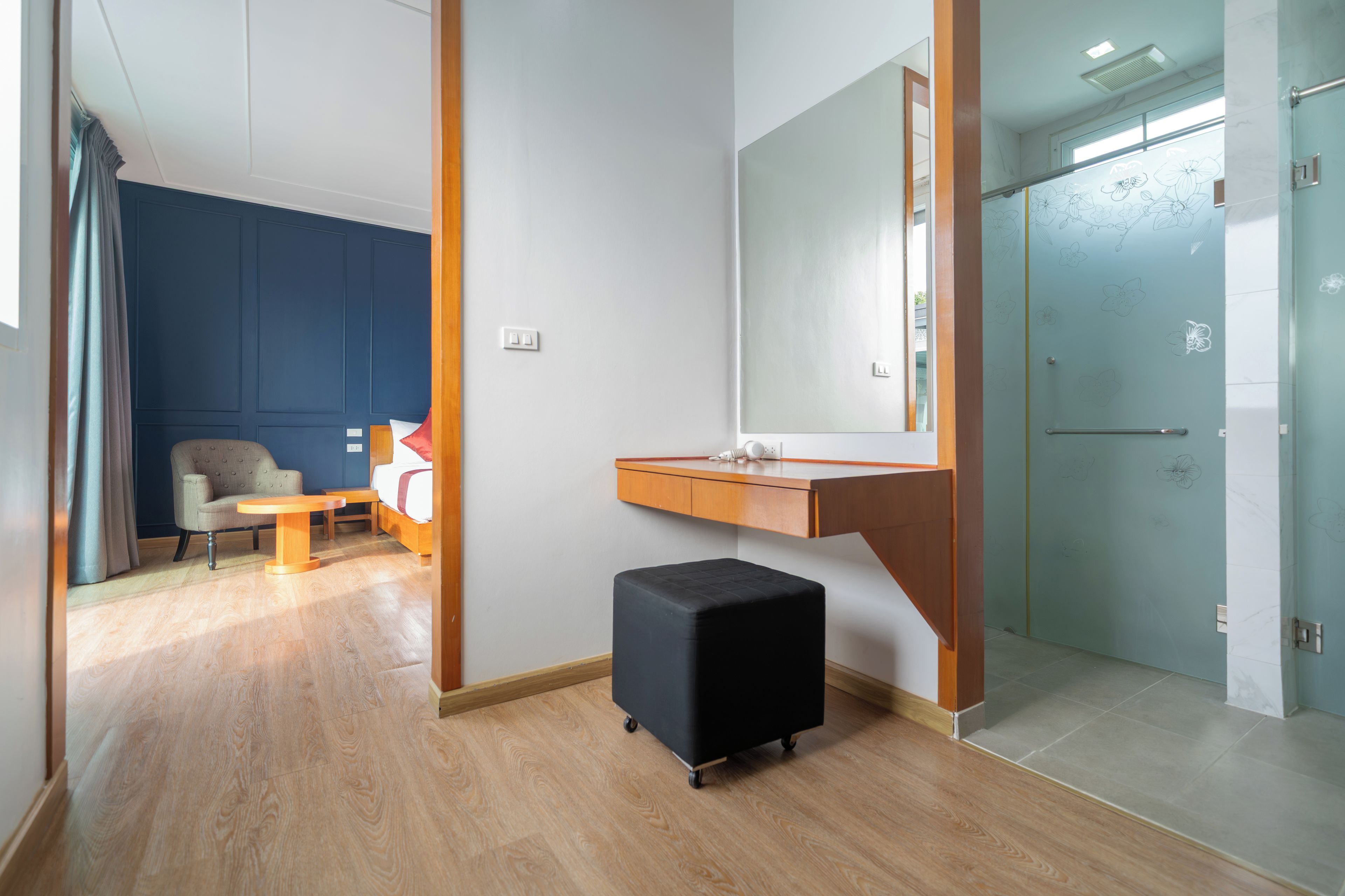 Een moderne hotelkamer met een houten vloer, een klein zwart poefje, een houten bureau, een spiegel en een glazen douchedeur.