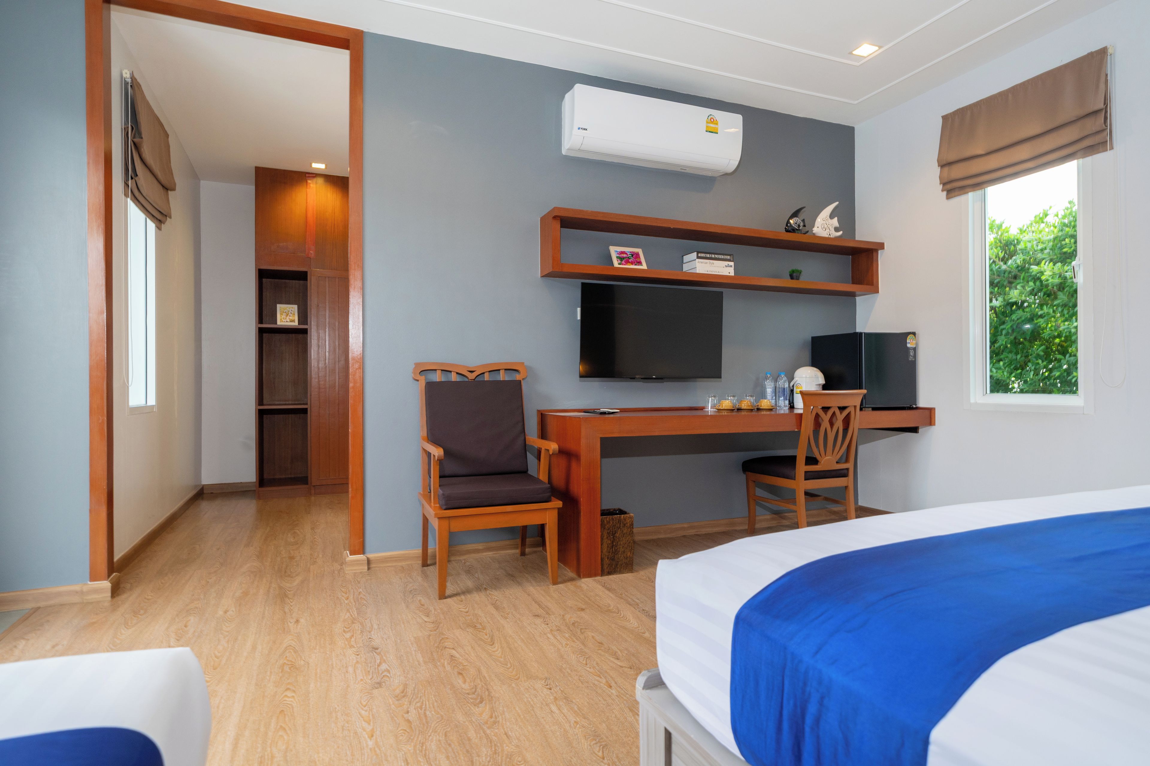 A hotel room with a bed, a desk with a chair, a television, and a window with a view of greenery.