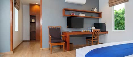A hotel room with a bed, a desk with a chair, a television, and a window with a view of greenery.
