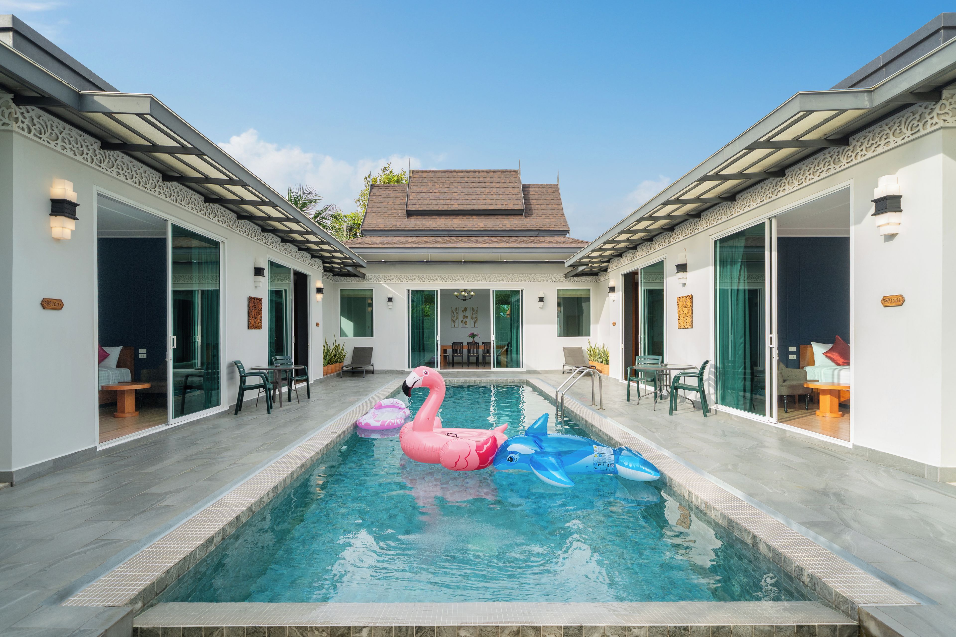 Una piscina con flotadores de plátano y delfín inflables, rodeada por una casa moderna con puertas de vidrio y un patio empedrado.