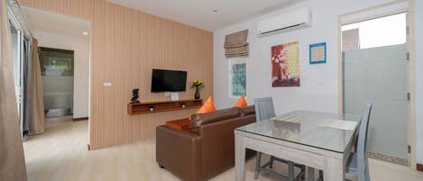 A modern living room with a brown leather sofa, a glass dining table, and a flat-screen TV mounted on the wall.
