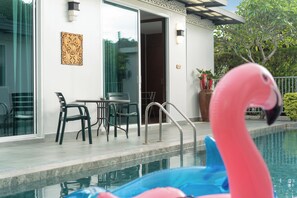 A swimming pool with a pink flamingo float, a patio with chairs and a table, and a building with a glass door and wall art.