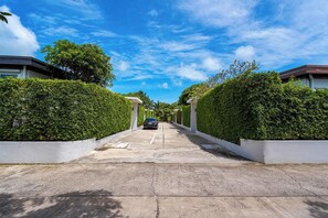 A residential street with a car parked in the middle, flanked by neatly trimmed hedges and white walls. - Phuket La Siesta Pool Villa And Pool Access (Rawai)