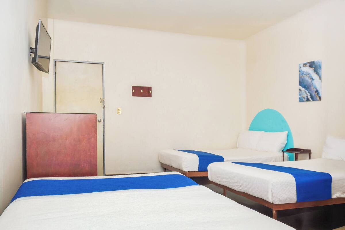 Une chambre d’hôtel avec trois lits simples, une armoire en bois, un téléviseur à écran plat fixé au mur et une petite table avec une chaise.