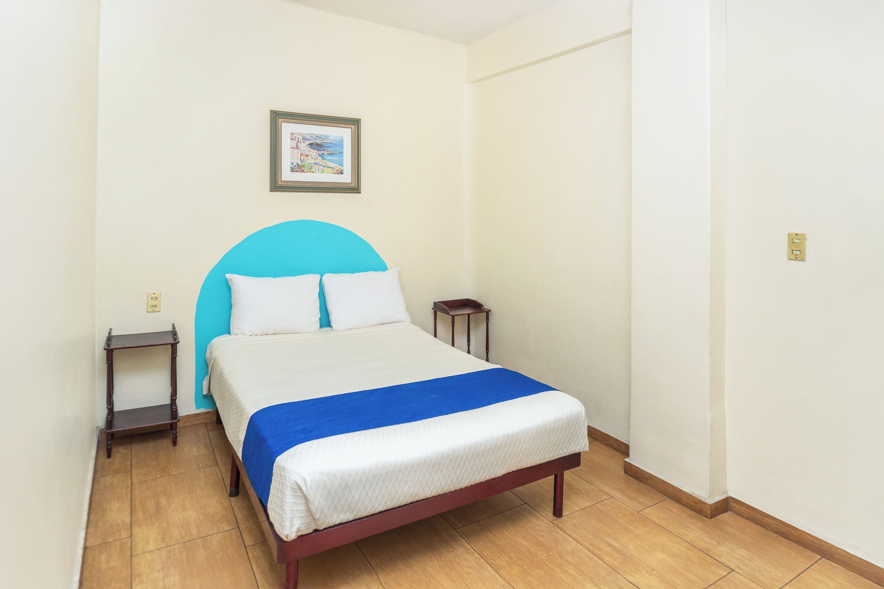 A neatly made bed with a blue headboard, a small bedside table, and a wall-mounted picture.
