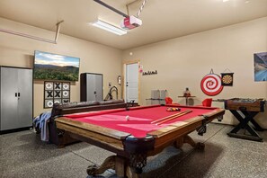 A pool table with a red felt surface, a foosball table, and a dartboard in a room with beige walls and a ceiling light.