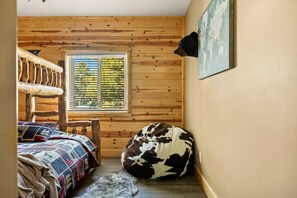 Une pièce chaleureuse avec un mur lambrissé en bois, un lit recouvert d’une couverture à motifs, un panier pour chien et une fenêtre avec des stores.