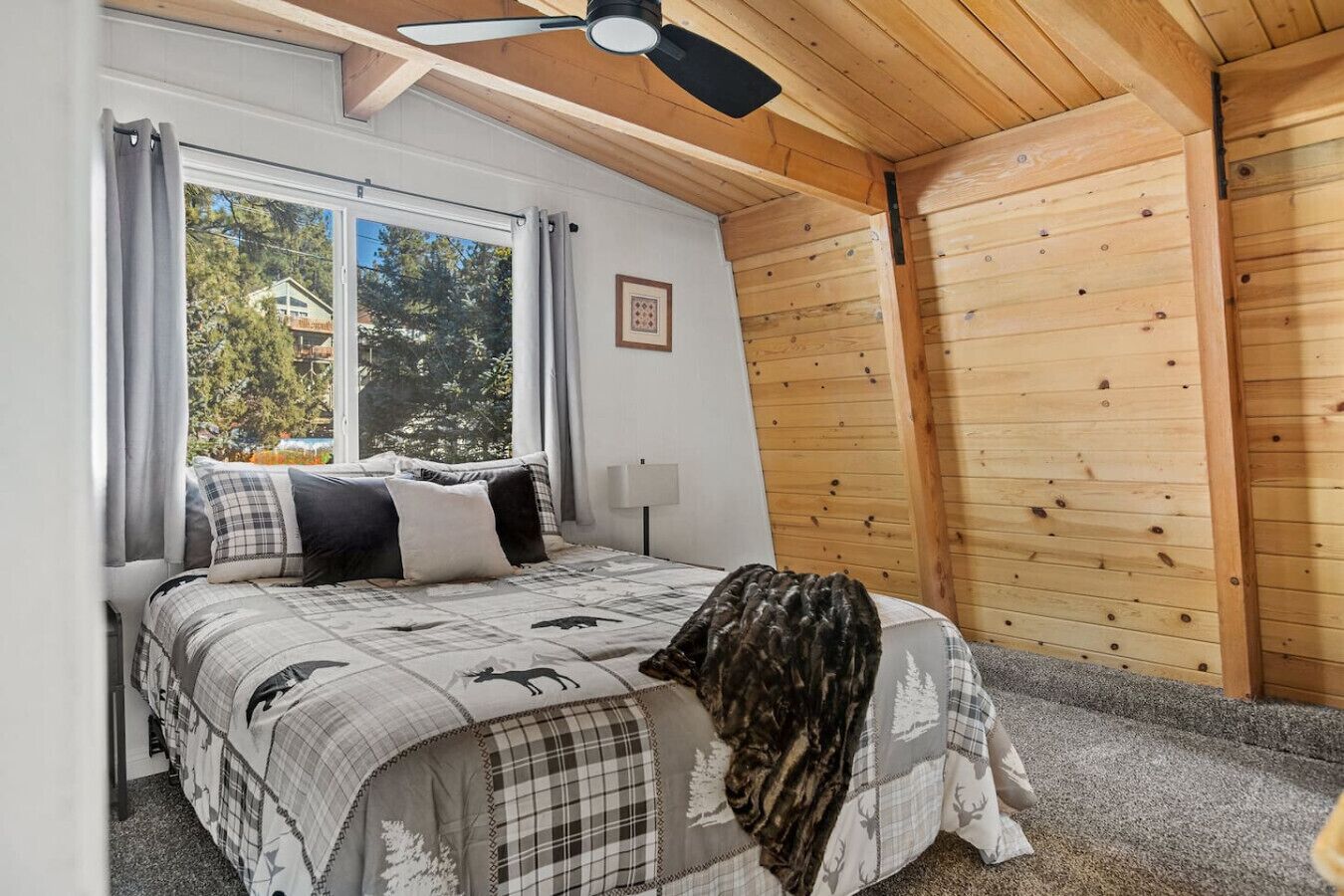 A bedroom with a bed, a ceiling fan, a window with curtains, and a view of trees and a house outside.