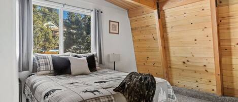 A bedroom with a bed, a ceiling fan, a window with curtains, and a view of trees and a house outside.