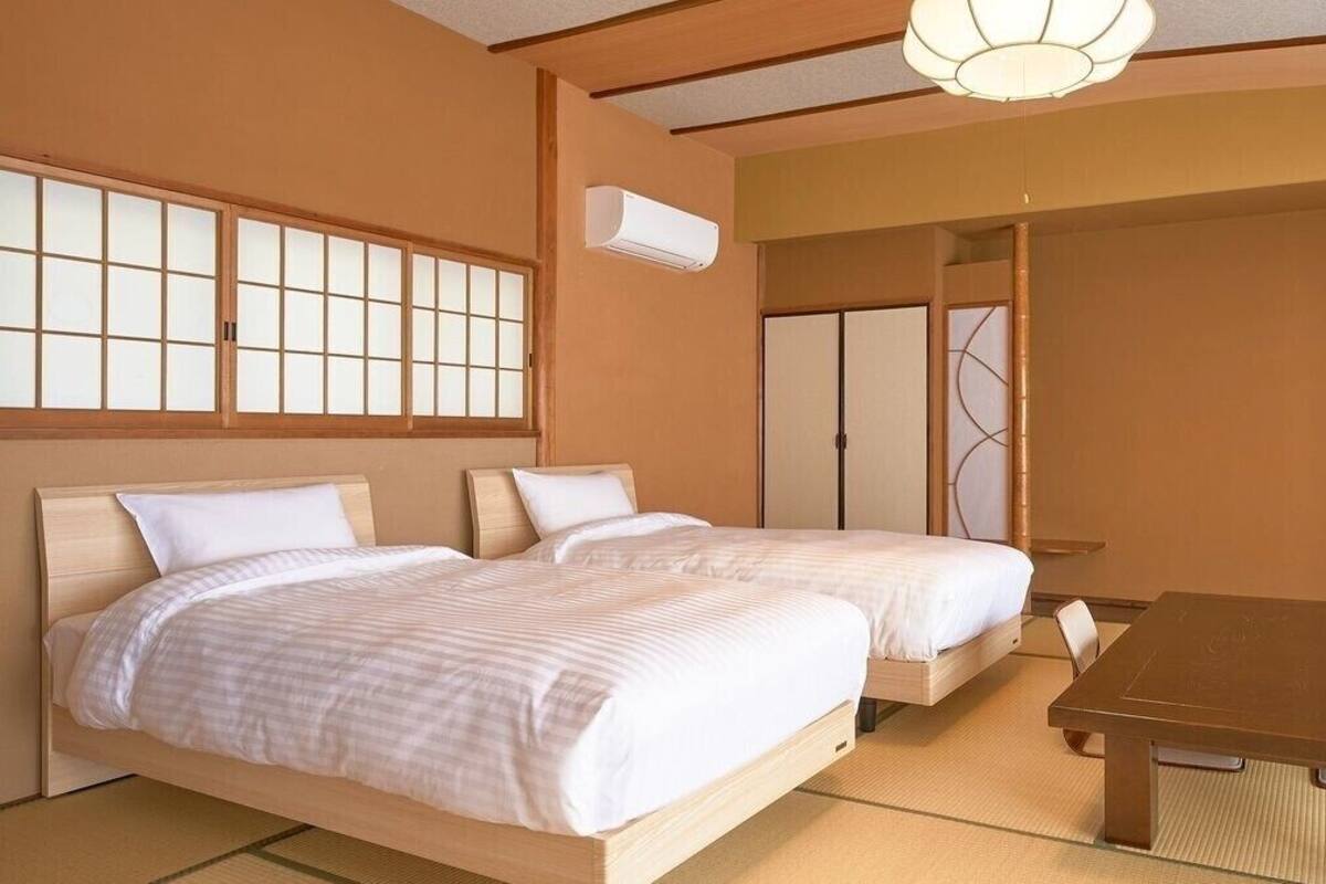 Una habitación tradicional japonesa con suelo de tatami, dos camas de madera con ropa de cama blanca, una mesa baja de madera y puertas correderas.