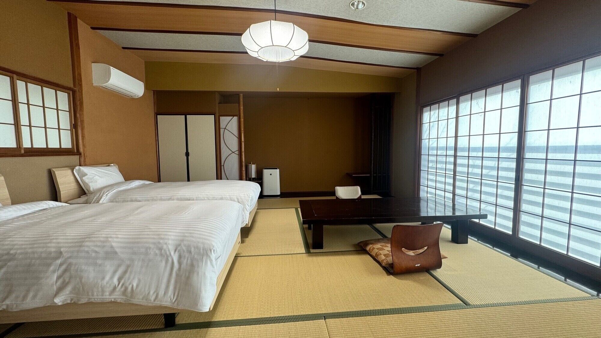 A hotel room with two beds, a desk, a chair, a large window, and a ceiling fan.