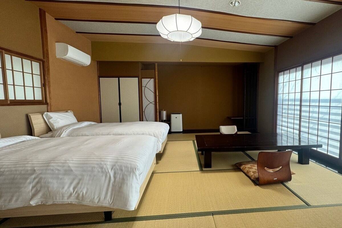 Habitación de hotel con dos camas, un escritorio, una silla, un ventanal grande y un ventilador de techo.