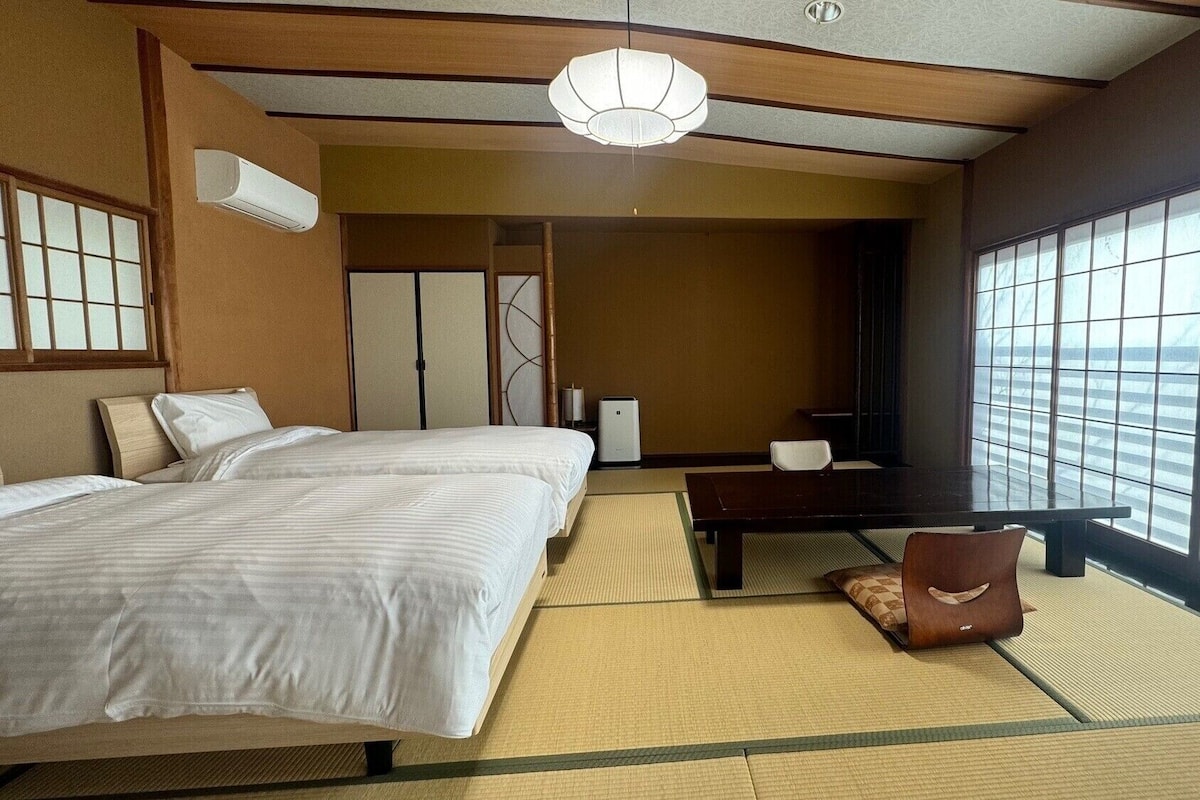 A hotel room with two beds, a desk, a chair, a large window, and a ceiling fan.