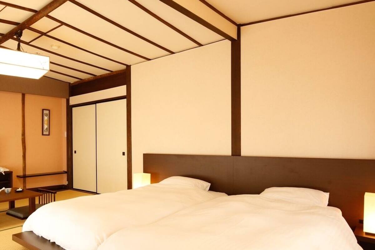 Habitación de hotel con una cama grande, cabecera de madera, suelo de tatami, mesita de noche y un muro al estilo tradicional japonés.