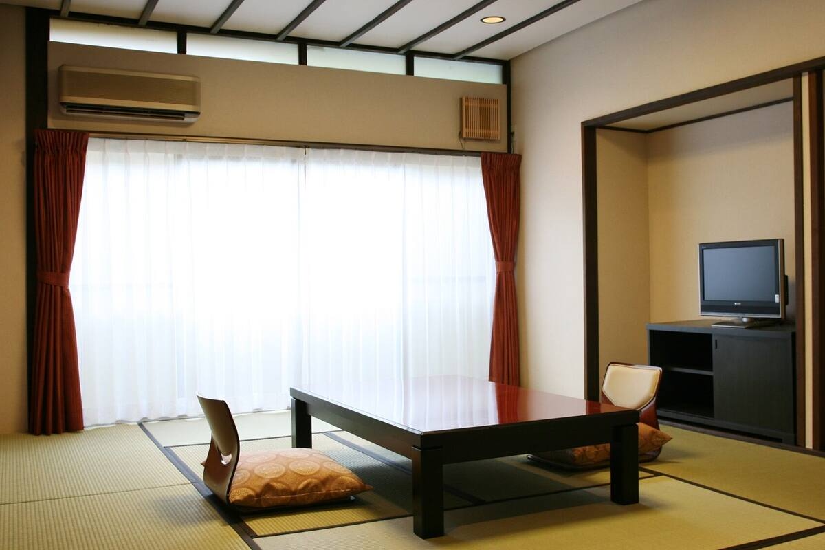 Una habitación japonesa tradicional con suelo de tatami, una mesa baja, una silla acolchada, un televisor sobre un mueble y puertas correderas.
