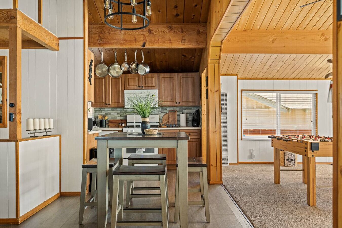 A cozy kitchen with wooden cabinets, a central island with chairs, hanging pots, and a foosball table.