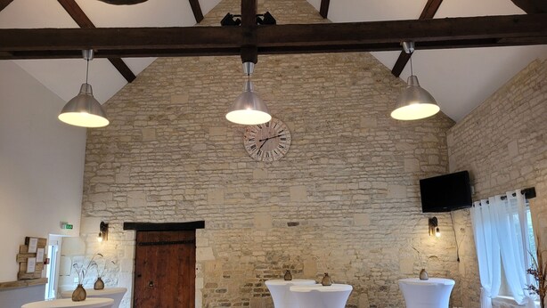 A rustic room with exposed stone walls, wooden beams, and a large clock. There are white tables and chairs, and a wooden door. - Magnificent farmhouse on an estate 5 minutes from Caen town center (LOUVIGNY)
