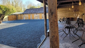 An outdoor seating area with tables and chairs, a gravel ground, and a wooden building with a sloped roof in the background. - Magnificent farmhouse on an estate 5 minutes from Caen town center (LOUVIGNY)