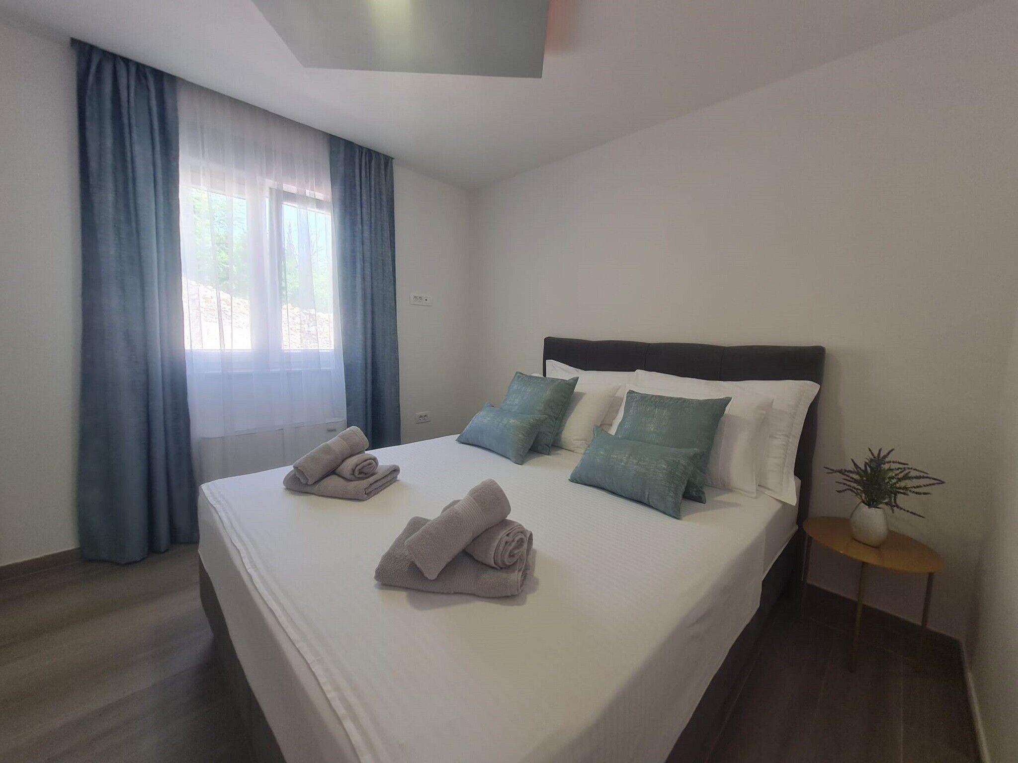 A neatly made bed with pillows and folded towels, a small side table with a plant, and a window with curtains.