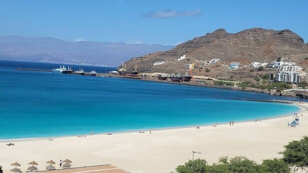 Escena costera con aguas azules cristalinas, una playa arenosa y montañas al fondo.