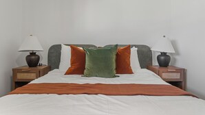 Een net opgemaakt bed met een wit laken, oranje en groene kussens en een bruine deken. Er zijn twee nachtkastjes met lampen en een klein lade.