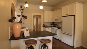 Une cuisine avec des meubles blancs, un plan de travail noir, deux tabourets en bois, un réfrigérateur blanc et une cuisinière.