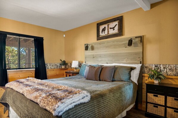 A bedroom with a large bed, a wooden headboard, a window with curtains, and a small bedside table with a lamp.