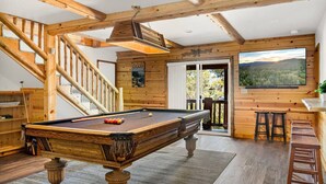 A wooden pool table in a room with a staircase, a bar area, and a large window with a view of a forest.