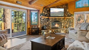 A cozy cabin interior with a stone fireplace, wooden beams, and a large window overlooking a forested area.