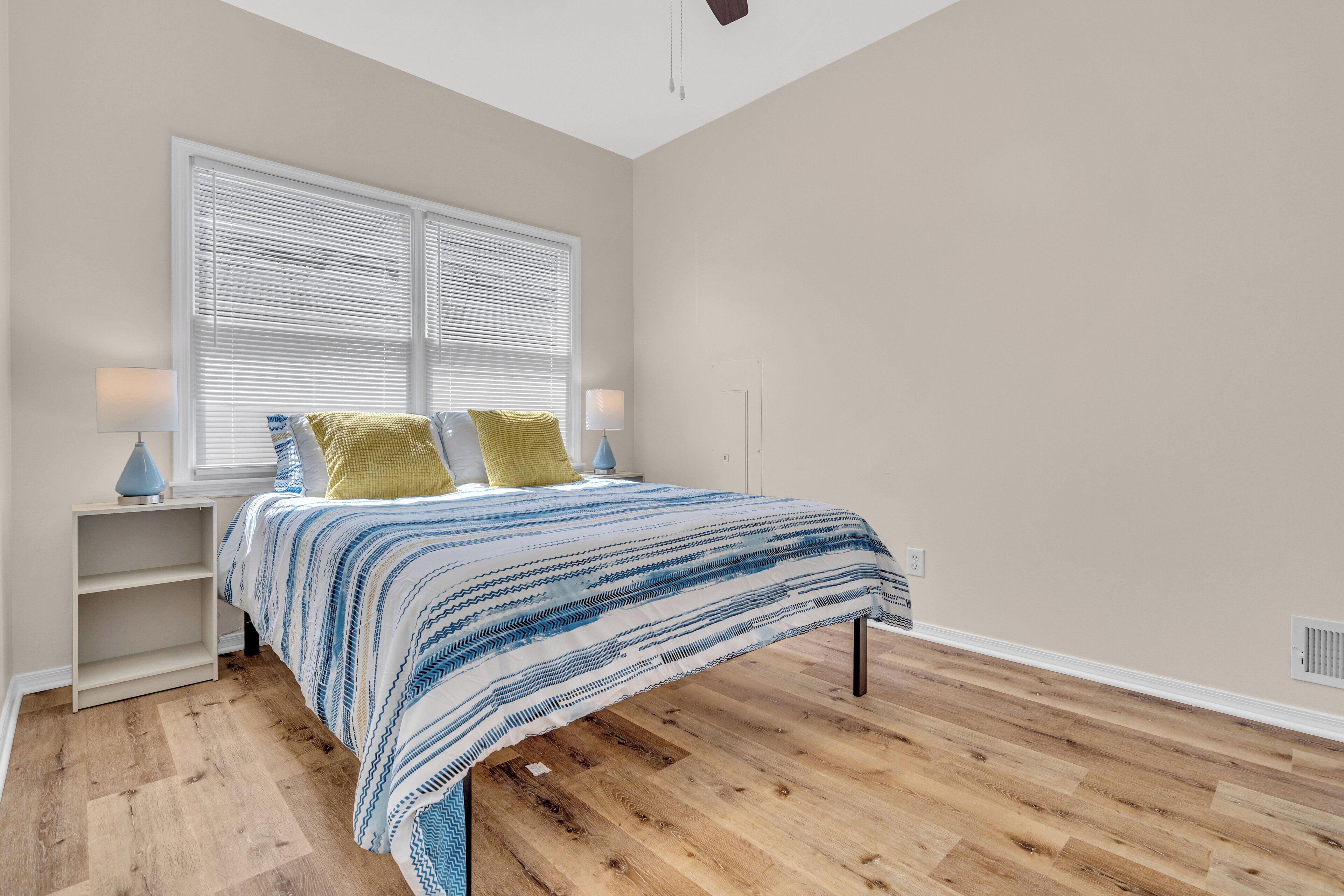 A bedroom with a bed, two bedside tables, a window with blinds, and a wooden floor.
