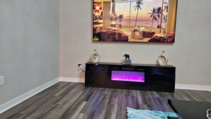 Een woonkamer met een grote flatscreen televisie aan de muur, een donkere tv-meubel eronder en een blauw, geribbeld vloerkleed op de houten vloer.