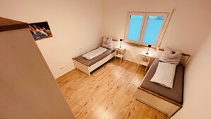 Un dormitorio pequeño y acogedor con dos camas individuales, una ventana con persianas, una mesita de noche con lámpara y un cuadro colgado en la pared.