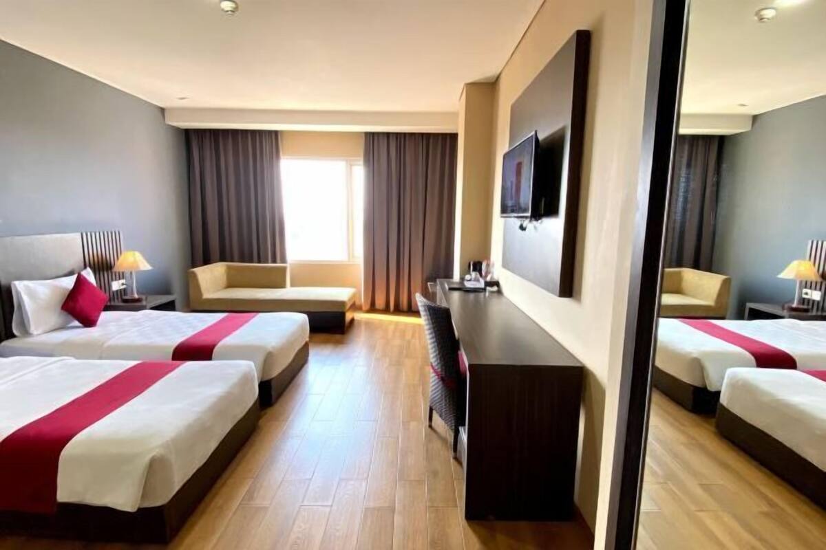 A hotel room with three beds, a desk, a TV, and a large window with curtains.