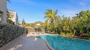 A swimming pool with a curved edge, surrounded by a tiled deck and lush greenery, including palm trees and a hedge.