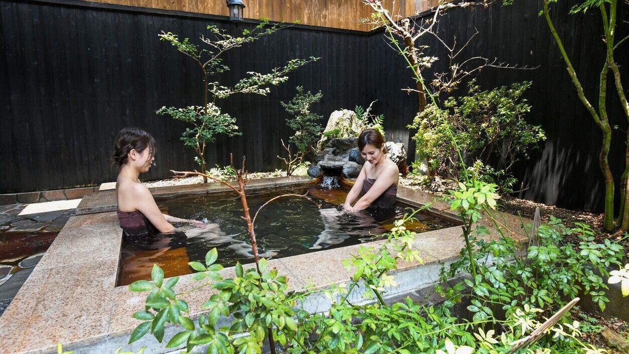 A small outdoor pool with two people sitting in it, surrounded by greenery and a wooden fence.