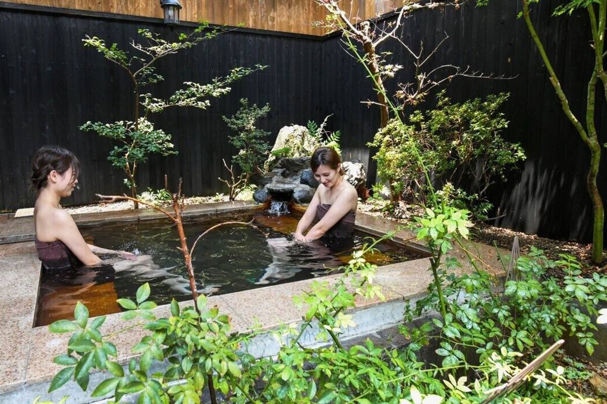 A small outdoor pool with two people sitting in it, surrounded by greenery and a wooden fence.