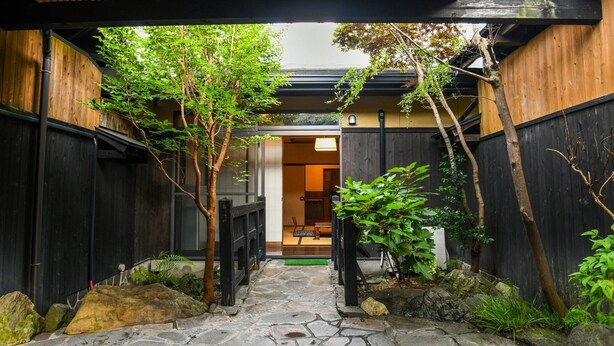 Ein gepflasterter Weg führt zu einem traditionellen japanischen Eingang mit Holzzäunen und Grünpflanzen.