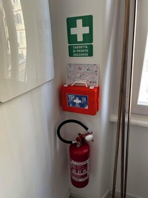 Ein an der Wand befestigtes Erste-Hilfe-Set, ein Feuerlöscher und ein Fenster mit Blick auf Gebäude.