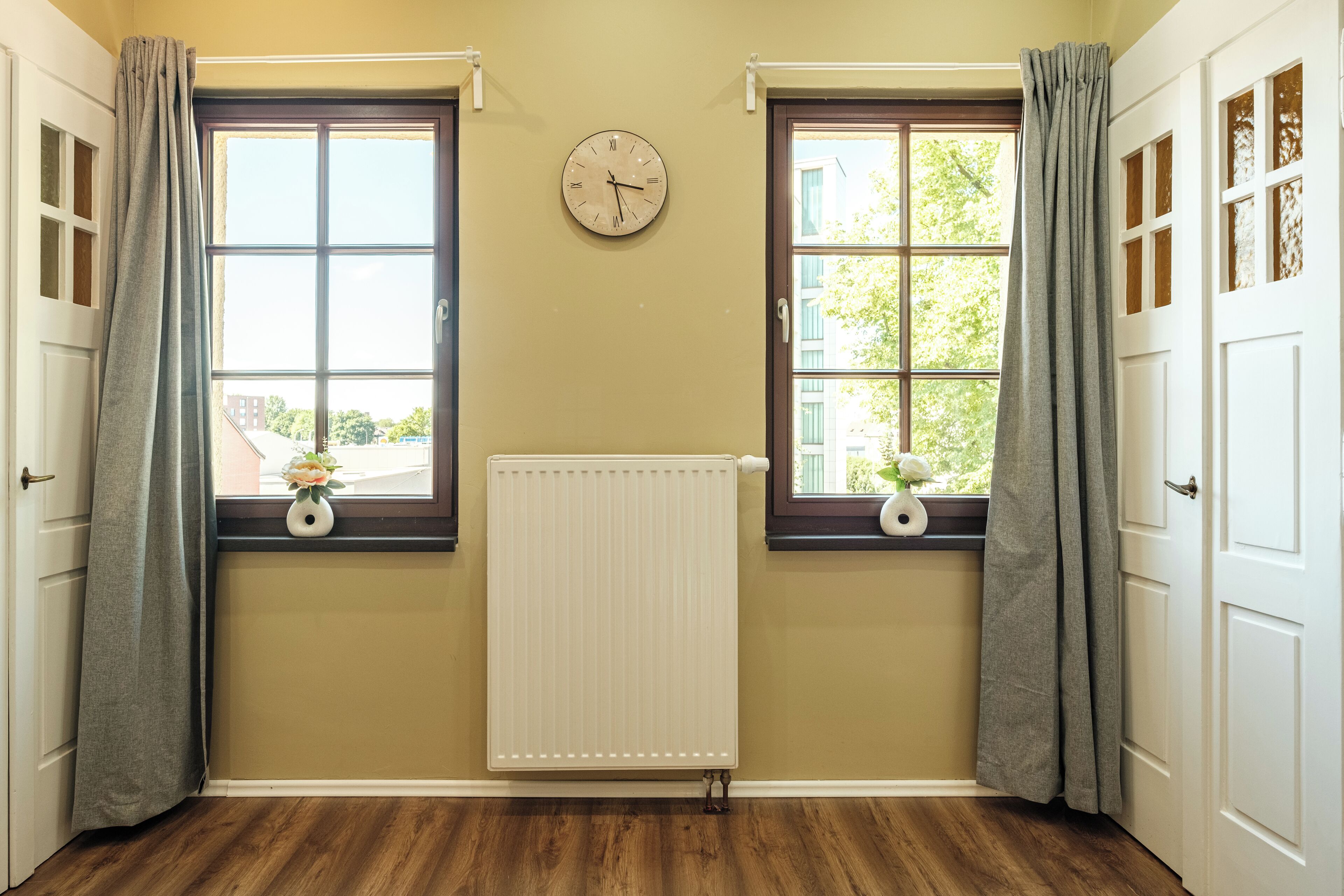 A room with two windows, a radiator, and a clock on the wall.