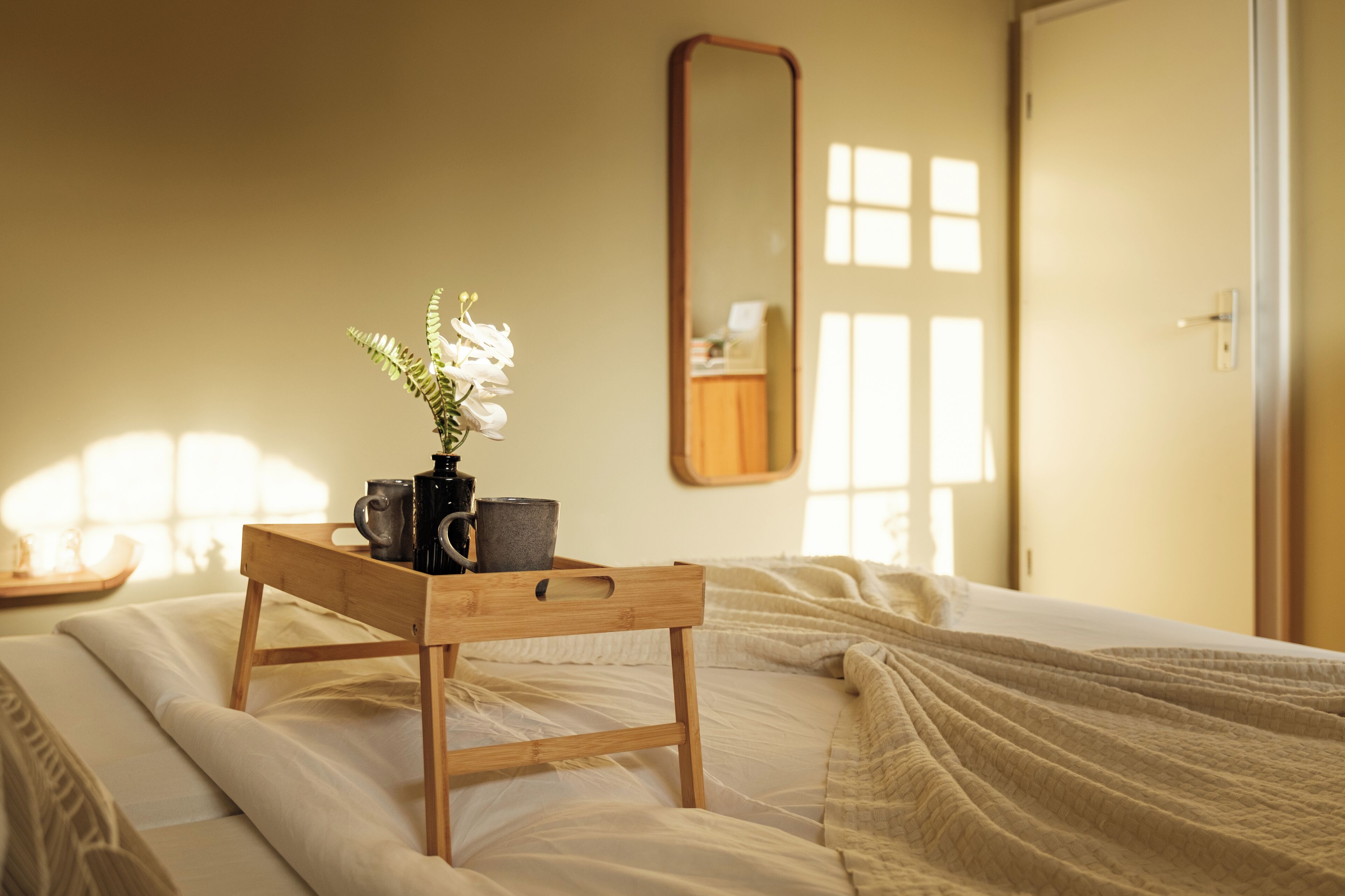 A neatly made bed with a wooden tray holding a vase of flowers and two mugs, a mirror on the wall, and a door with a window.