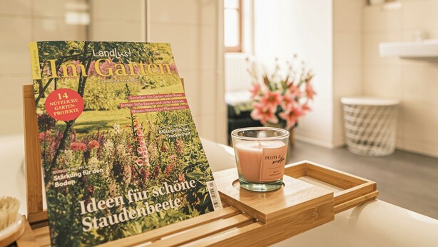 Eine Zeitschrift mit dem Titel „Landlust Im Garten“ auf einer Holzschale mit einer Kerze daneben in einem Badezimmer.
