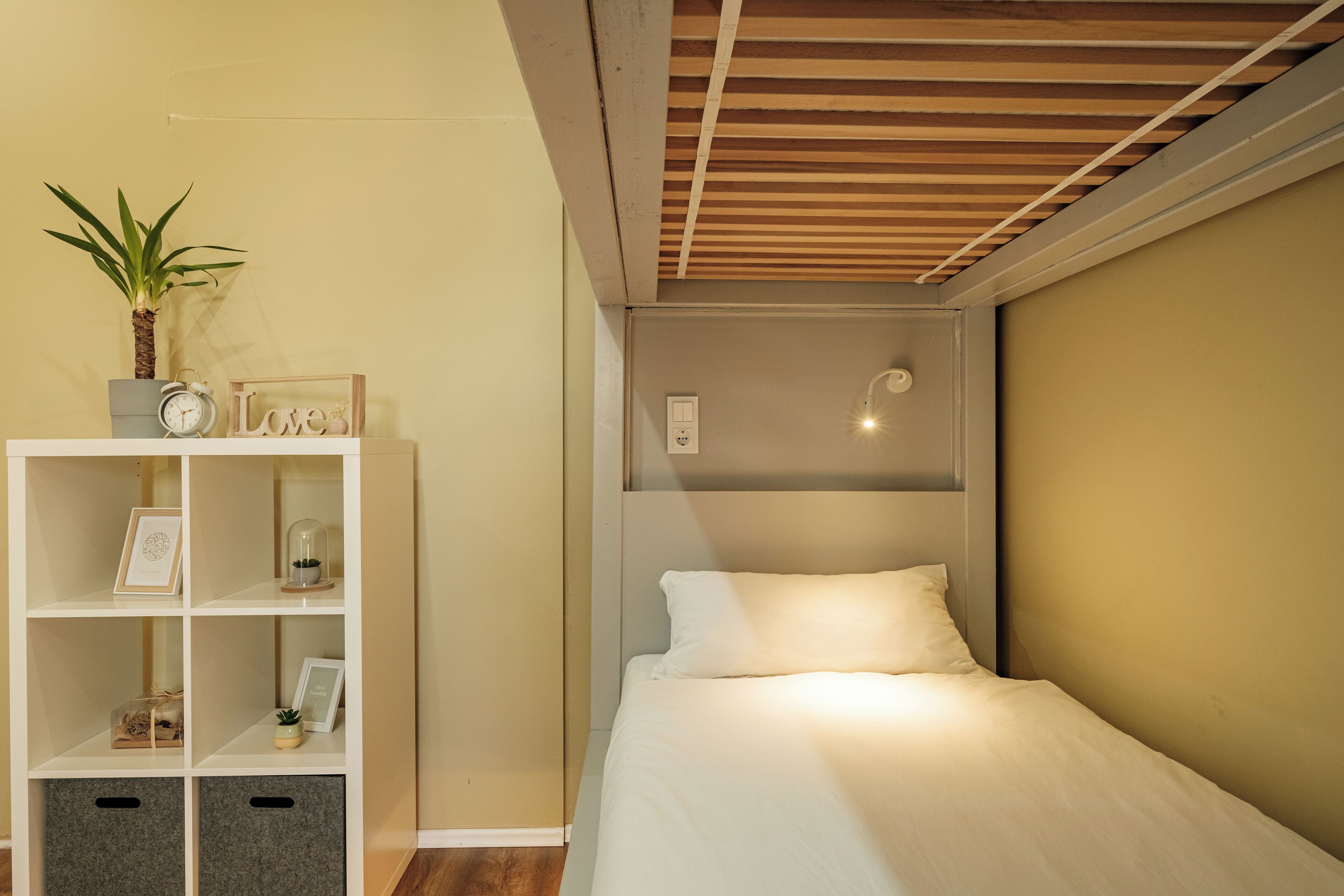 Une chambre à coucher avec un lit, une étagère blanche contenant divers objets, une plante en pot et un plafond avec des poutres en bois.