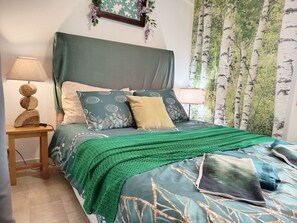 A bed with a green blanket, patterned pillows, and a headboard. There is a nightstand with a lamp and a decorative item. - L’Écrin des Bois - Au bord de l'Aude (Espéraza)