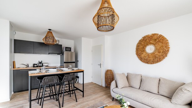 Eine moderne Küche mit dunklen Schränken, einem Holztisch und schwarzen Barhocker. Ein beiges Sofa und ein runder Couchtisch mit Blumenvase ergänzen das Bild.