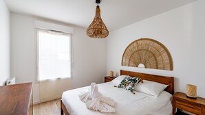 Ein ordentlich bezogenes Bett mit weißer Daunendecke, ein Nachttisch aus Holz mit einer gewobenen Lampe und ein großes, gewebtes Wandbild.