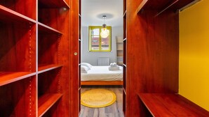 A narrow walk-through closet with wooden shelves, a bed with white linens, a yellow rug, and a hanging light fixture.