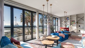 Un amplio espacio de estar con ventanas grandes, un sofá azul, una mesa de centro de madera y una estantería.