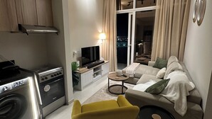 A modern living room with a sofa, a yellow armchair, a coffee table, and a TV. There is a kitchen area with a stove and a washing machine. The room has a large window with curtains and a view of the city at night. - Cozy Golf & Lake View 1-bedroom apartment in Dubai,Damac Hills (Dubai)